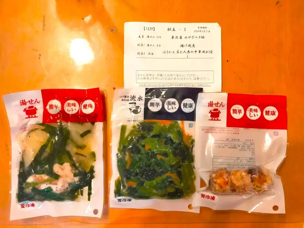 わんまいるの健幸ディナー。湯せんや流水で調理できる個包装の冷凍惣菜セットで、豚肉とニラの煮物、揚げ焼売、ほうれん草と人参の中華風お浸しが並んでいる。