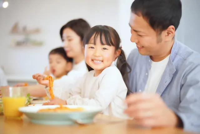 家族で食卓を囲み、笑顔で食事をする様子。父親と娘が座りごはんを食べている。