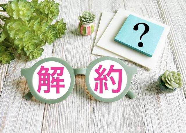 解約という文字が書かれた丸いプレートをメガネのレンズのように並べたイメージと、背景に多肉植物と疑問符の付いた付箋が置かれている写真。